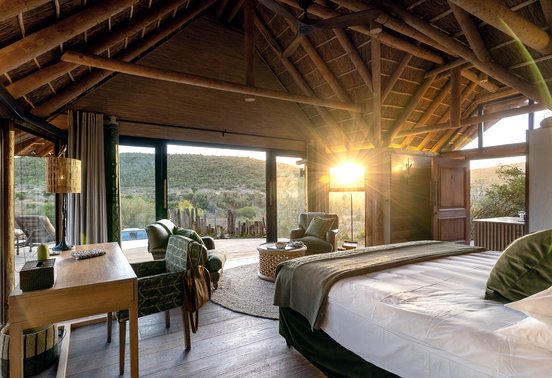 Luxury thatched bedroom with king bed and golden sunrise light flooding through glass doors at Kwandwe Great Fish River Lodge