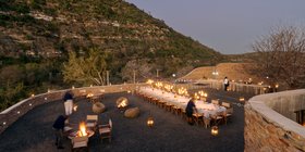 Outdoor boma dinner at night with long candlelit table, fire pits and mountain backdrop at Kwandwe Great Fish River Lodge