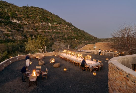 Outdoor boma dinner at night with long candlelit table, fire pits and mountain backdrop at Kwandwe Great Fish River Lodge
