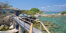 Stone chalet perched above turquoise waters connected by wooden walkway at Kaya Mawa, Likoma Island.