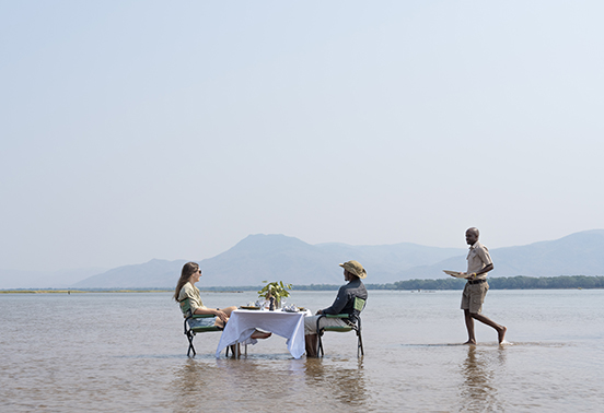 Guests dining at a white-clothed table set in the shallow waters of the Zambezi River with a waiter serving lunch, Lower Zambezi National Park.