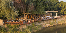 Elevated view of Sausage Tree Camp’s riverfront deck and lounge shaded by trees along the Zambezi River, Zambia.