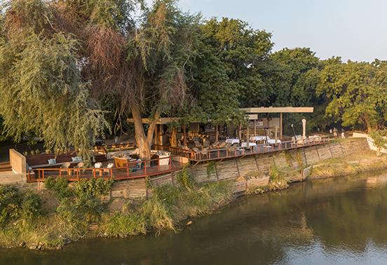 Elevated view of Sausage Tree Camp’s riverfront deck and lounge shaded by trees along the Zambezi River, Zambia.