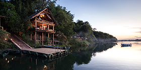 Riverfront house at Tongabezi Lodge overlooking the Zambezi River at sunset, with a private jetty below.