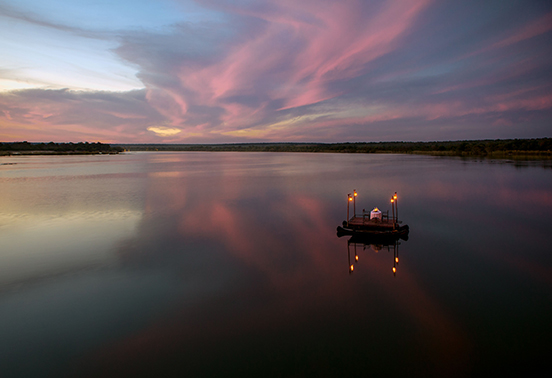 Private floating dinner platform illuminated by lanterns on the Zambezi River at sunset at Tongabezi Lodge, Zambia.