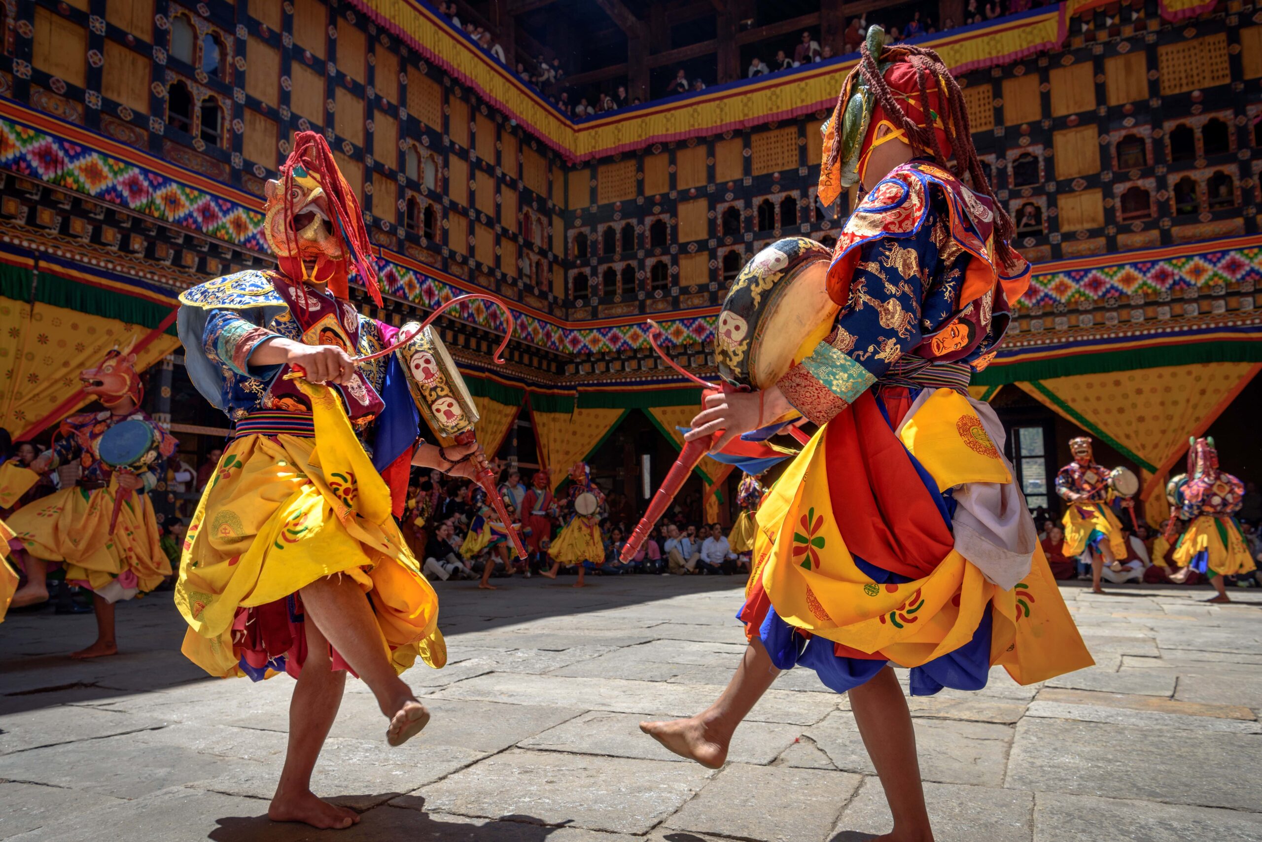 Performers in vibrant yellow and blue costumes dance barefoot during a colorful Buddhist festival at a monastery courtyard in Bhutan.