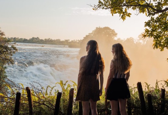 Two guests standing at the edge of Victoria Falls in golden mist, Zimbabwe