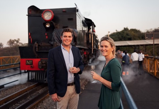 Smiling couple with champagne standing in front of the Bushtracks Express steam locomotive at Victoria Falls, Zimbabwe