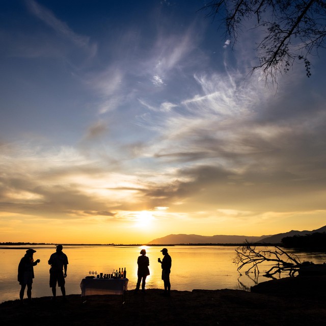 Silhouettes of guests at a sundowner table on the Zambezi River bank beneath a dramatic golden African sky, Lower Zambezi, Zambia