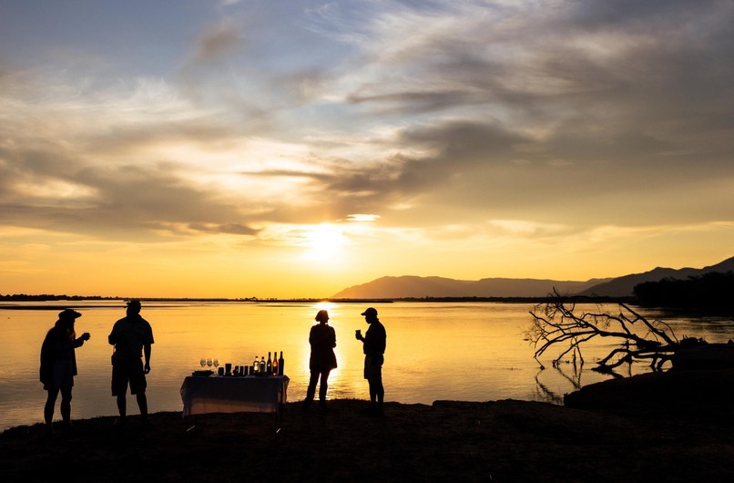 Silhouettes of guests at a sundowner table on the Zambezi River bank beneath a dramatic golden African sky, Lower Zambezi, Zambia