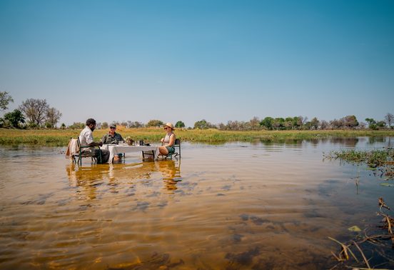 Guide and two guests dining at a table set in the shallows of the Okavango Delta under an open blue sky, Botswana