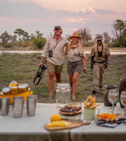 Two laughing guests and a guide walking toward a sundowner table set with drinks and snacks at dusk in the Okavango Delta, Botswana