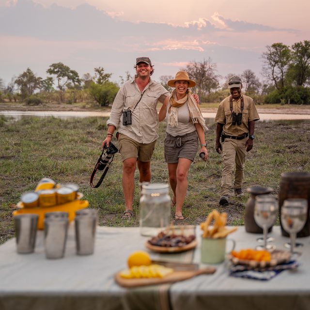 Two laughing guests and a guide walking toward a sundowner table set with drinks and snacks at dusk in the Okavango Delta, Botswana