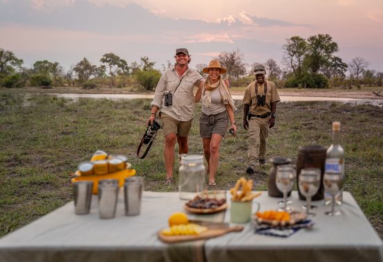 Two guests and a guide walking toward a bush sundowner table set with food and drinks at sunset in the Okavango Delta, Botswana