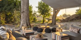 Open-sided camp lounge with striped ottomans and lanterns overlooking the Okavango Delta floodplains at sunset, Kweene Camp, Botswana