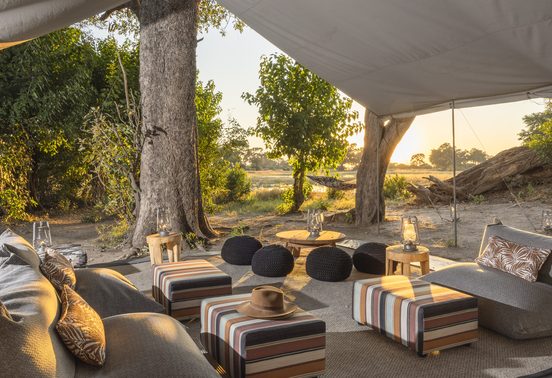 Open-sided camp lounge with striped ottomans and lanterns overlooking the Okavango Delta floodplains at sunset, Kweene Camp, Botswana