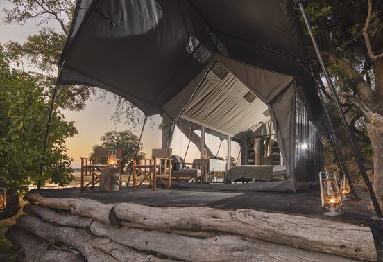 Safari tent exterior at dusk with lanterns glowing on a log deck overlooking the Okavango Delta at Kweene Camp, Botswana