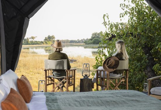 Two guests seated in camp chairs looking out over the Okavango Delta floodplains from inside their safari tent at Kweene Camp, Botswana