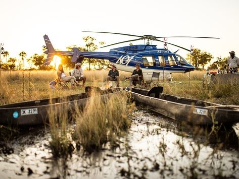 Guests gathered around a campfire beside a helicopter and traditional mokoros at sunset in the Okavango Delta floodplains, Botswana