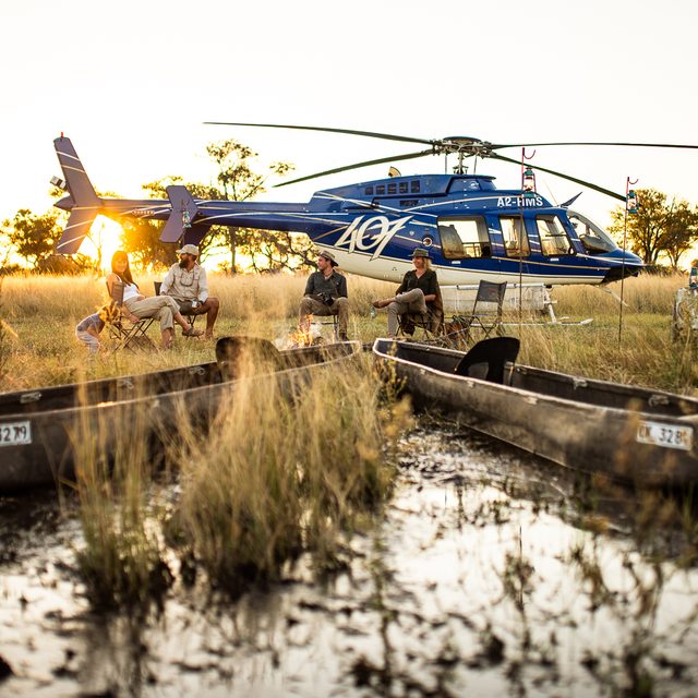 Guests gathered around a campfire beside a helicopter and traditional mokoros at sunset in the Okavango Delta floodplains, Botswana