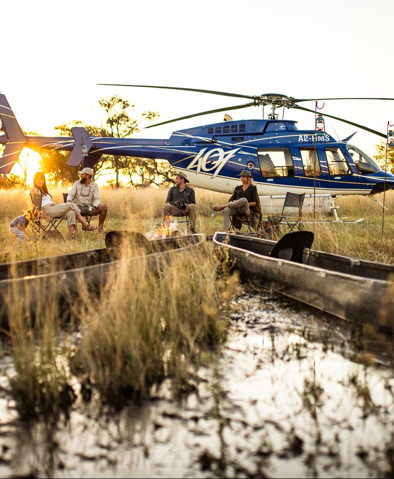 Guests gathered around a campfire beside a helicopter and traditional mokoros at sunset in the Okavango Delta floodplains, Botswana