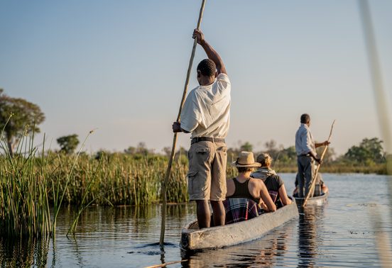 A poler standing and guiding a traditional mokoro through reed-lined waterways of the Okavango Delta with guests on board, Botswana