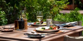 Outdoor dining table set with fresh food, white wine, and artisan tableware in dappled light at Karangoma, Okavango Delta, Botswana