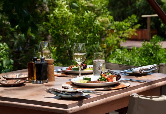Outdoor dining table set with fresh food, white wine, and artisan tableware in dappled light at Karangoma, Okavango Delta, Botswana