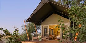 Safari tent exterior with timber deck and warm interior light glowing at dusk, framed by lush trees at Karangoma, Okavango Delta, Botswana