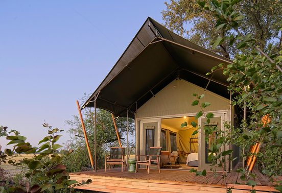 Safari tent exterior with timber deck and warm interior light glowing at dusk, framed by lush trees at Karangoma, Okavango Delta, Botswana