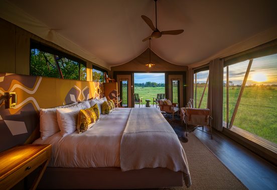 Luxurious safari tent interior with bold patterned headboard and king bed looking out over a sunset floodplain at Karangoma, Okavango Delta, Botswana